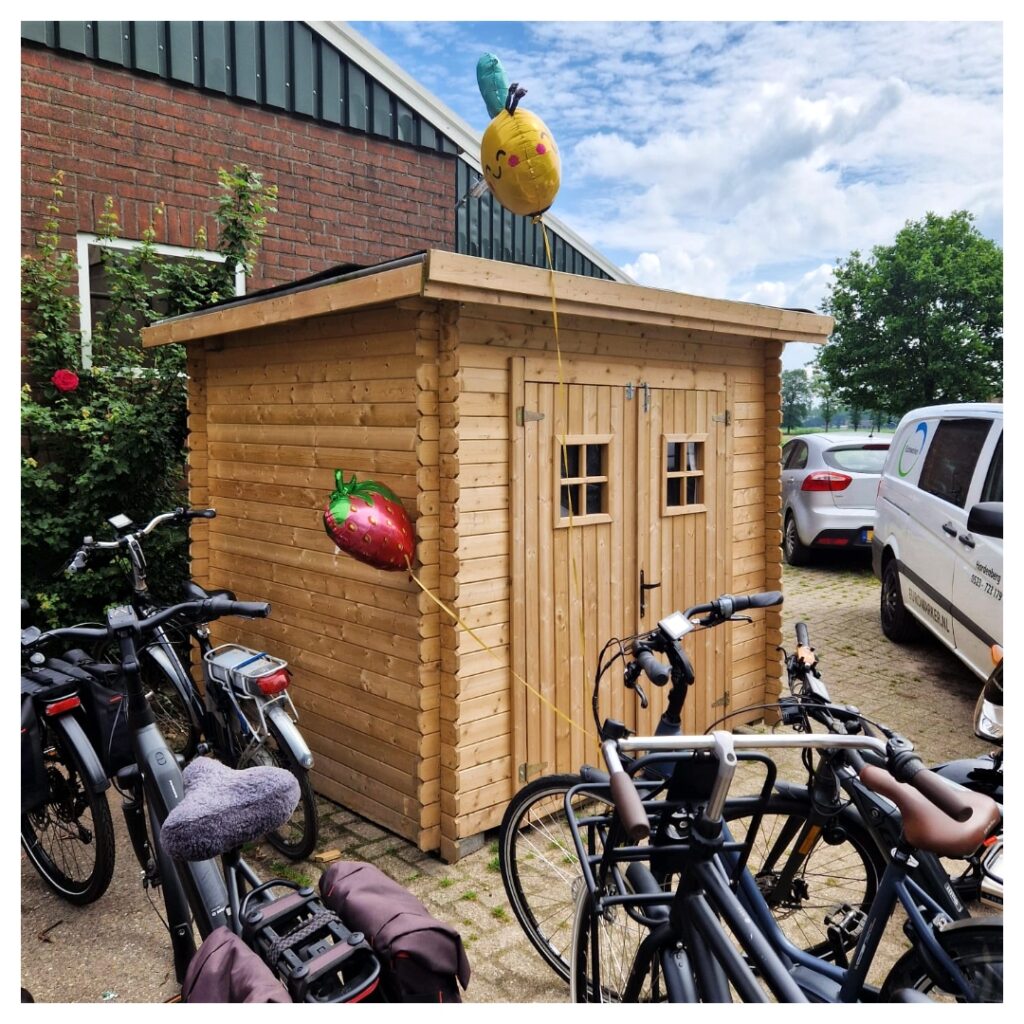 Een houten verkoophok met twee kleine ramen, versierd met ballonnen in de vorm van een aardbei en een ananas, omringd door fietsen op een oprit.