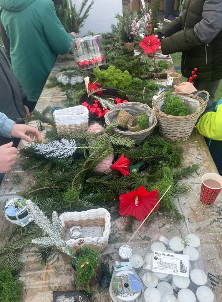 Tafel met kerstdecoratiematerialen zoals dennentakken, bloemen, en versieringen, terwijl mensen bezig zijn met het maken van kerstdecoraties.