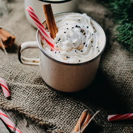 Een mok met slagroom, een kaneelstokje en een candy cane, omringd door kaneelstokjes op een jute ondergrond.